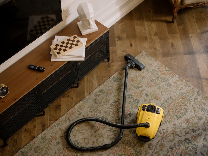 A person is using a yellow vacuum cleaner with a black hose attachment to deep clean a traditional patterned area rug in a domestic living room setting. The rug features beige, green, and reddish-oran
